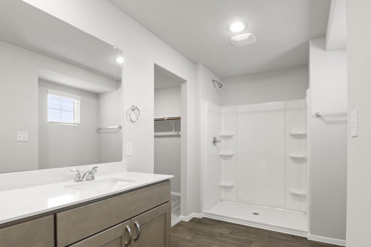 Image of a primary bathroom with a white vinyl walk-in shower, a connecting walk-in closet, and a white vanity with a large mirror