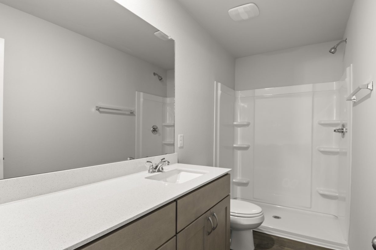 Image of a primary bathroom with brown cabinets, a white vanity and a large mirror