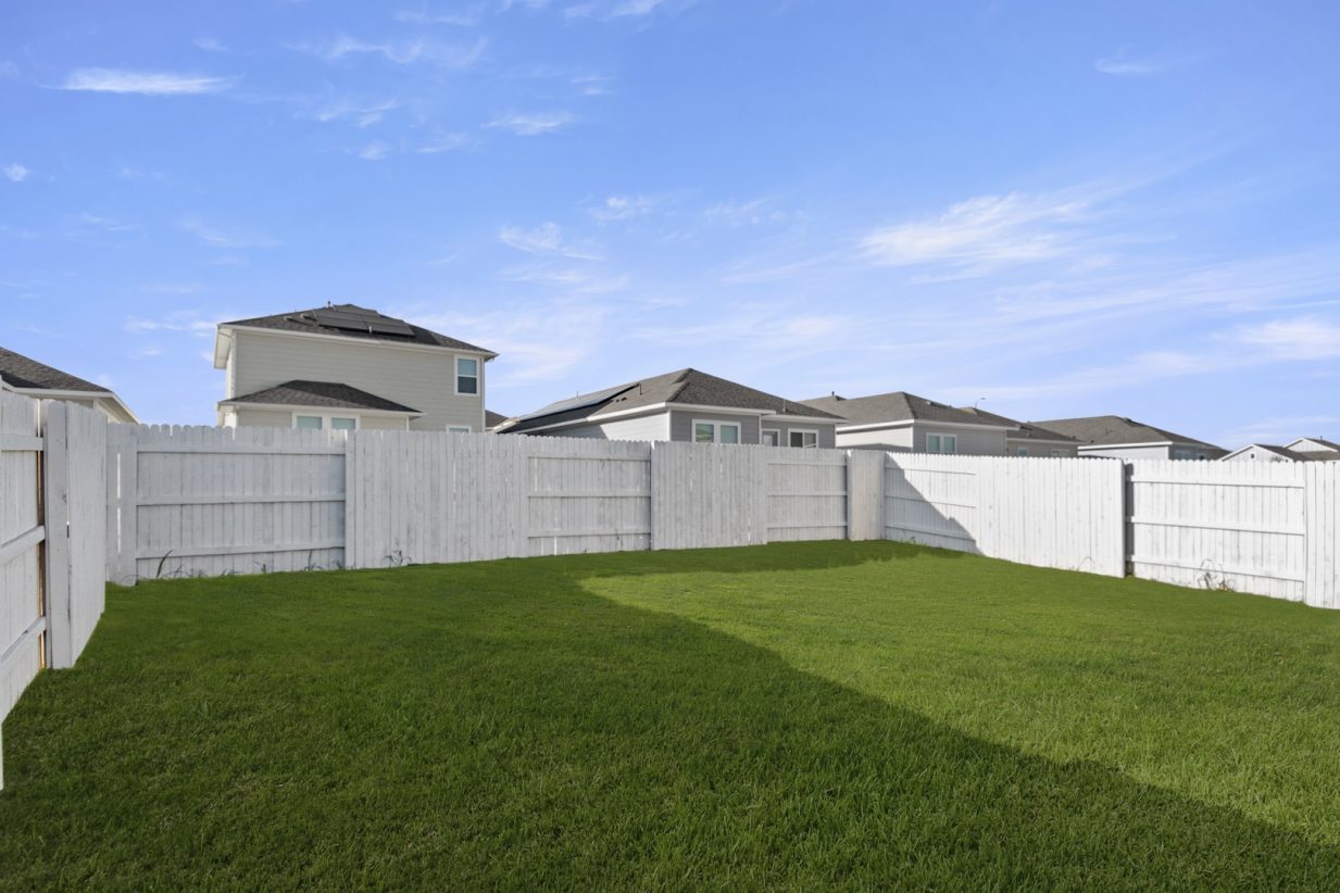Image of one story home backyard with green grass and a white wooden fence with a blue sky