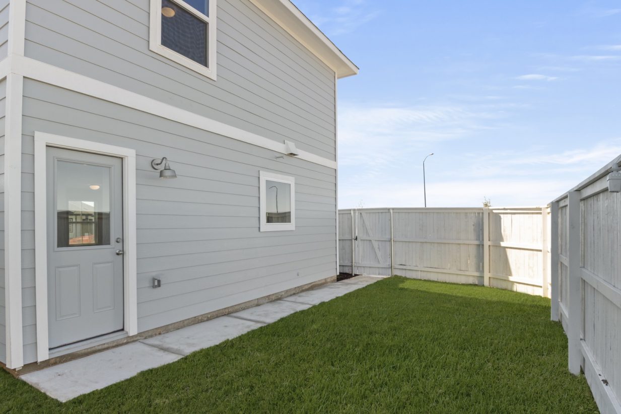 Image of a cottage home backyard with a backdoor, a white fence, cement walkway, and a blue sky