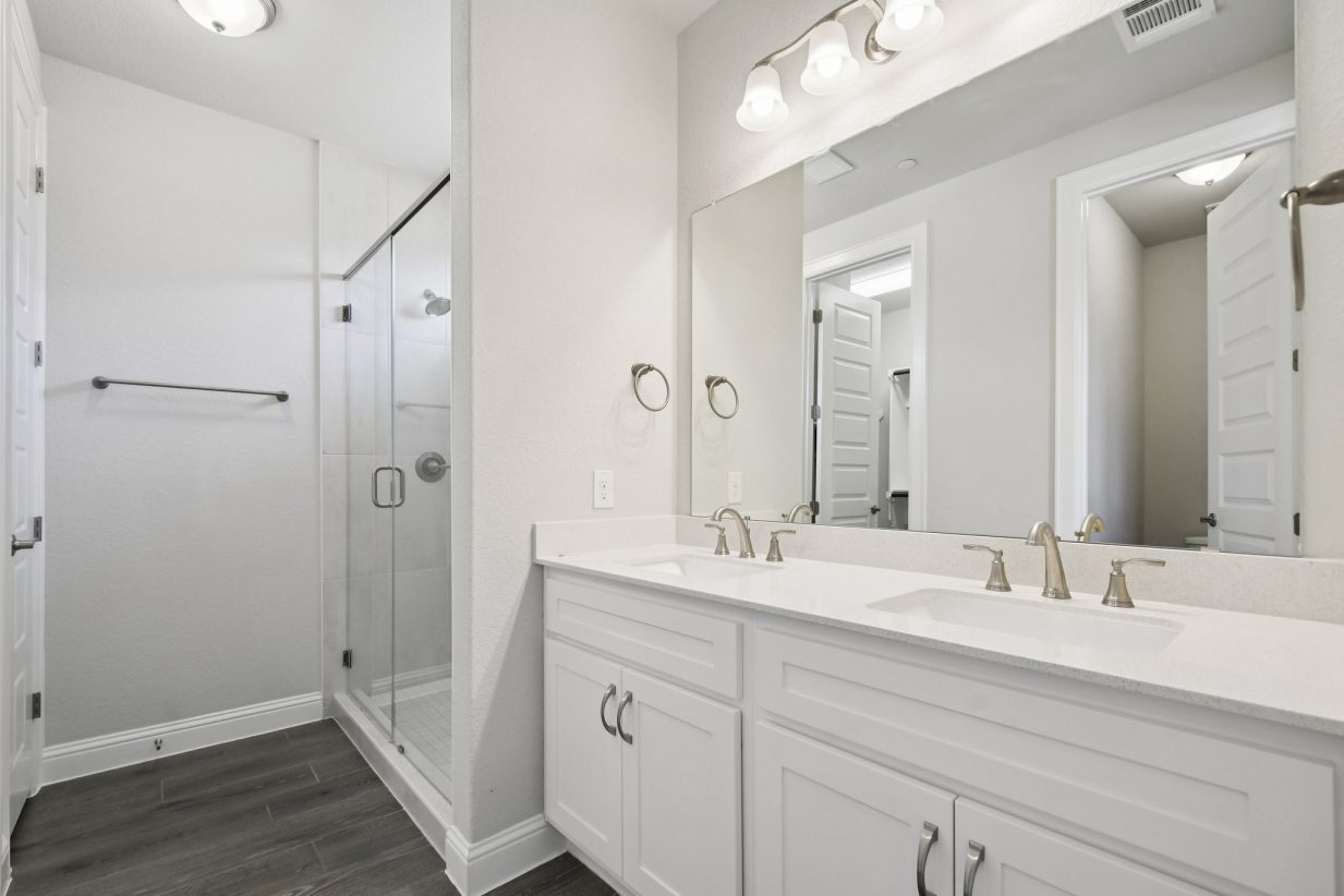 Image of a primary bathroom with a white jack and jill vanity, a large mirror, white cabinets and a walk-in shower