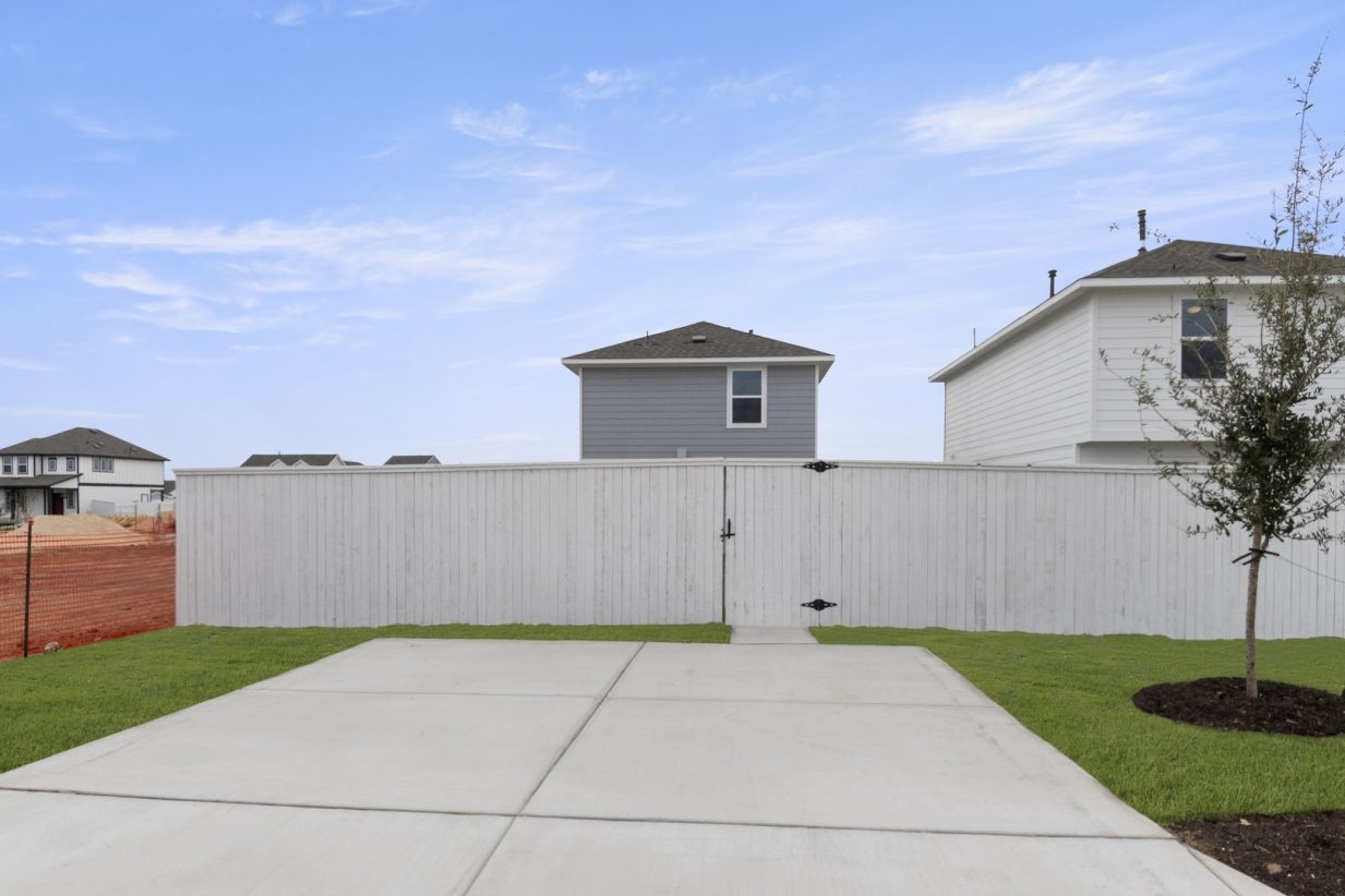 Image of a back cement driveway to a grey cottage two story home with a white fence