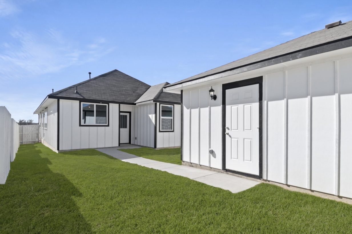 Image of a white one story house back exterior with a back white garage, green grass and a blue sky in the background