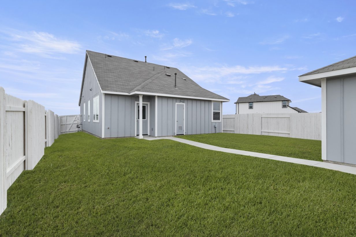 Image of a grey one story home back exterior with white trim, a green grass backyard, a white fence and a grey shed