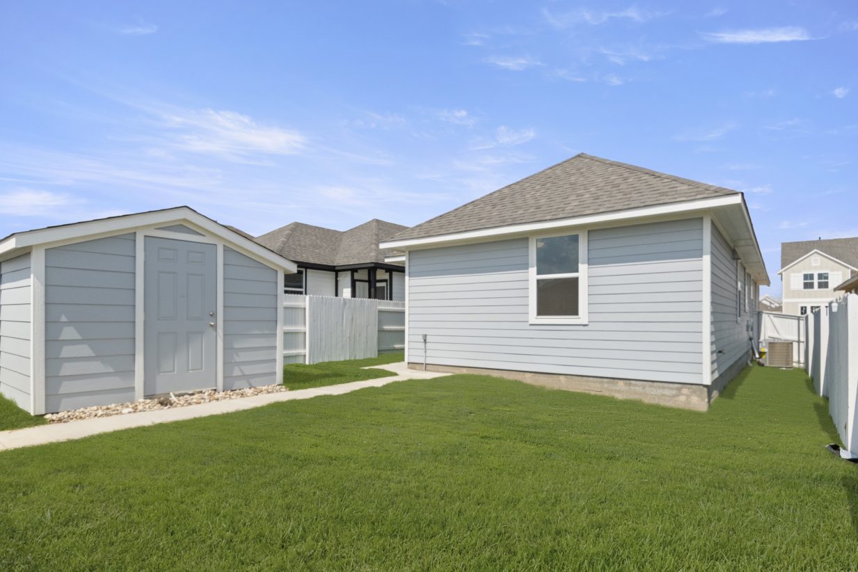 Image of back exterior of a blue one story home with a blue shed, green grass and a blue sky in the background