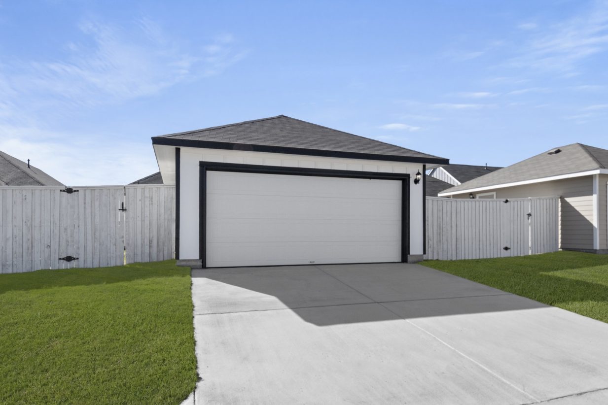 Image of an ally access garage with black trim, a cement driveway, green grass, white wooden fence and a blue sky in the background