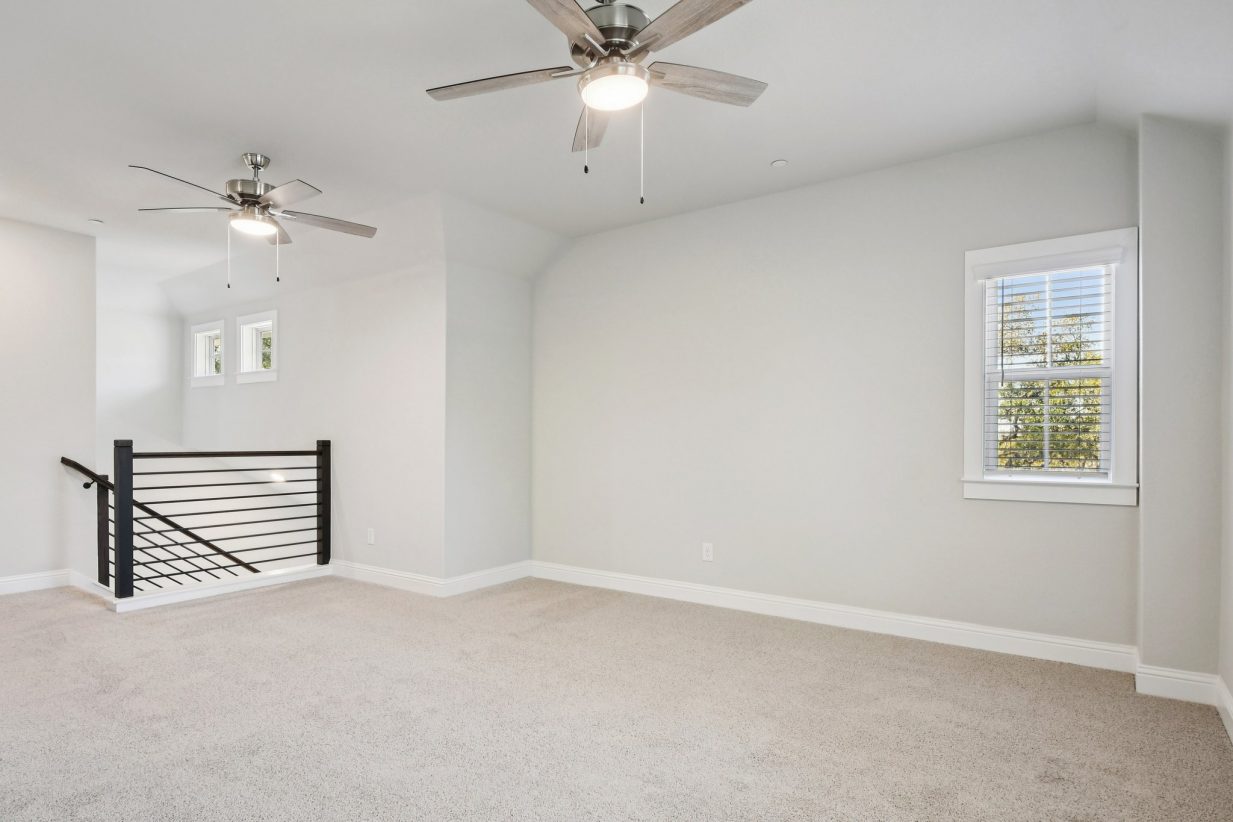 Image of an upstairs gameroom with light grey walls, tan carpeting, a black stair rail, and two ceiling fans