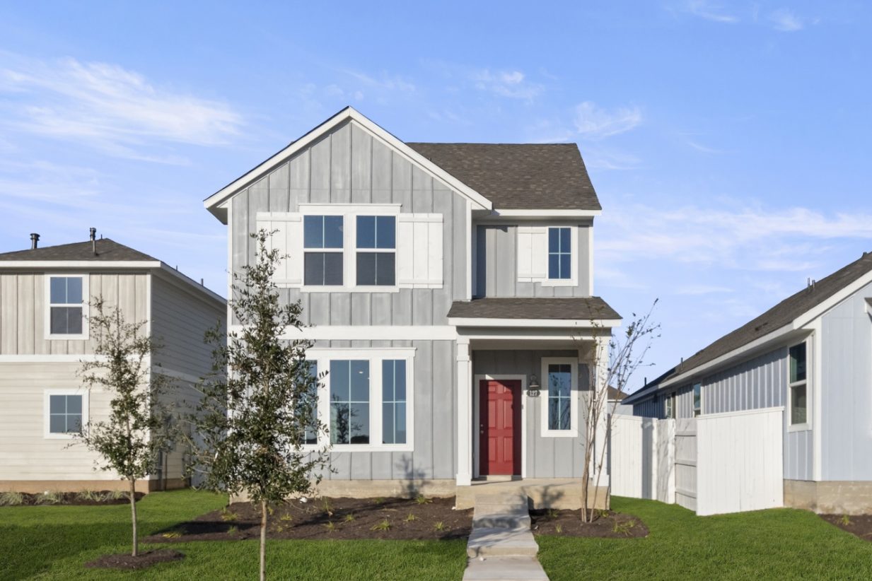 Image of the front exterior of a grey two story house with a red front door and white trim and shutters with green grass and a blue sky