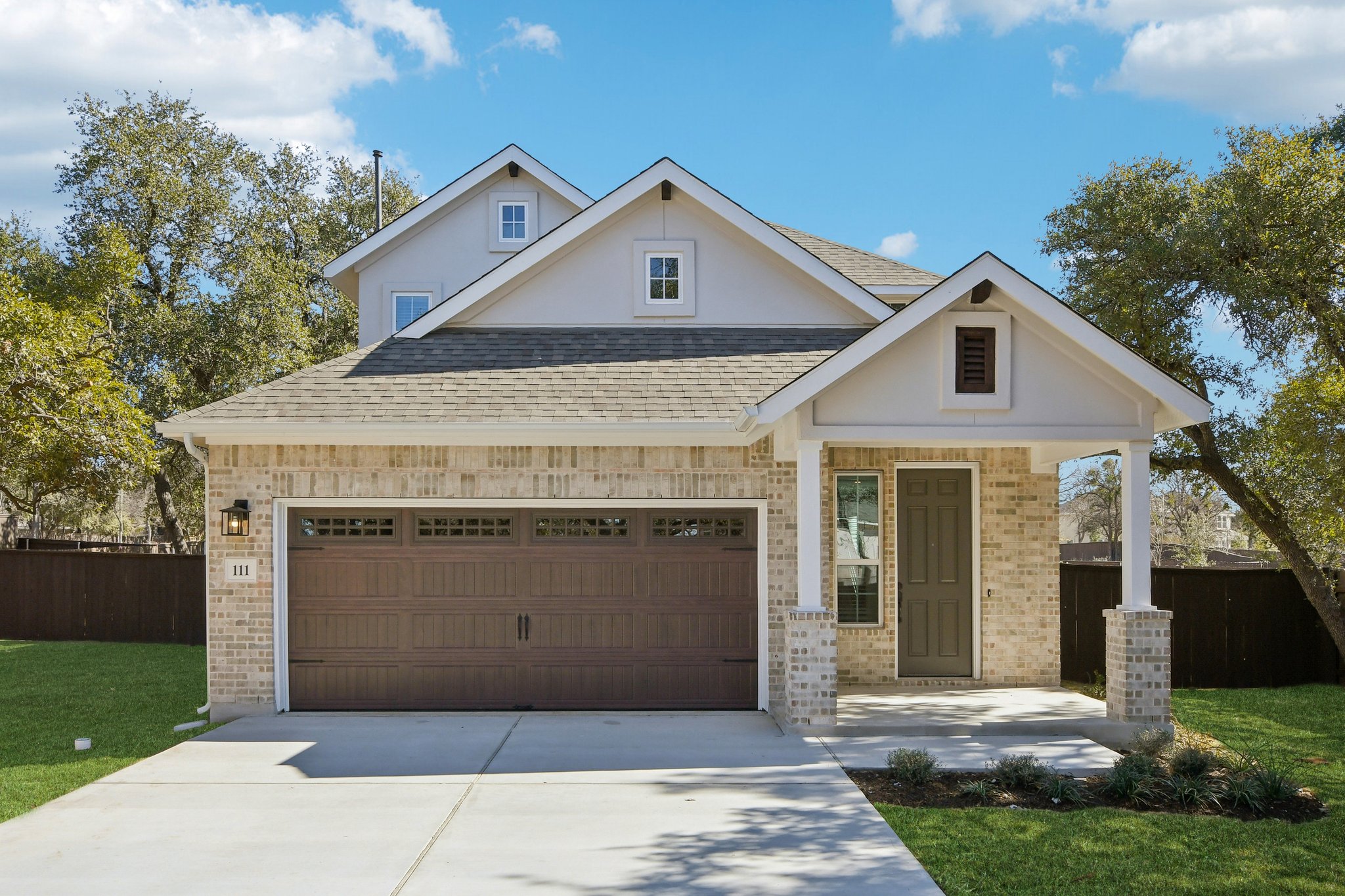 Image of a brick two story house with a brown garage door and front door, beige brick, green grass, and a blue sky in the background