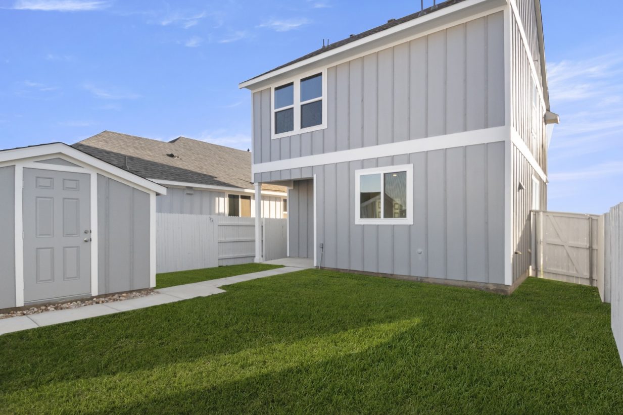 Image of the back exterior of a grey two story home with green grass and a shed, a white fence, and a blue sky