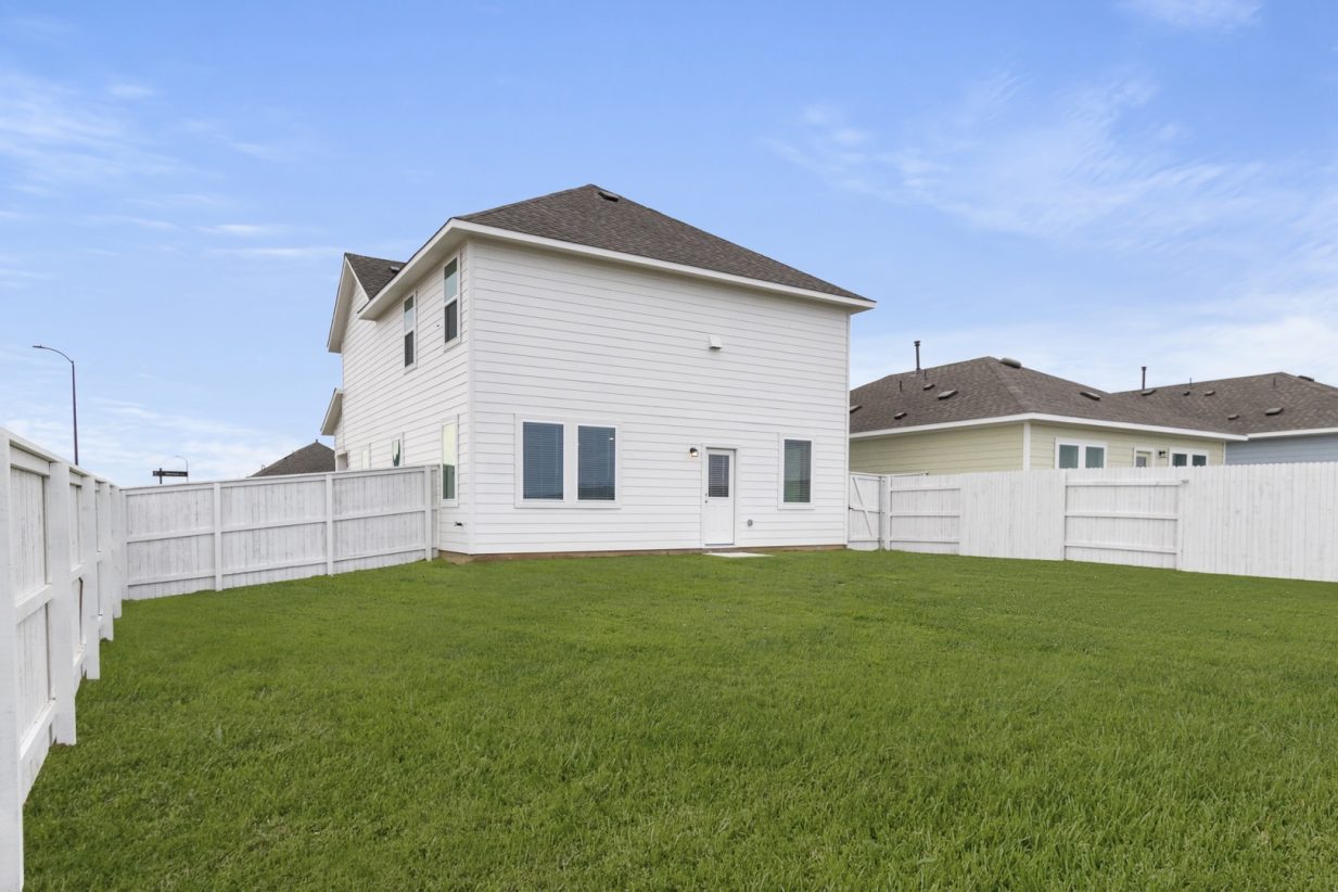 Image of the back exterior of a white and blue two story house with a large green grass yard, white wooden fence, and a clear blue sky