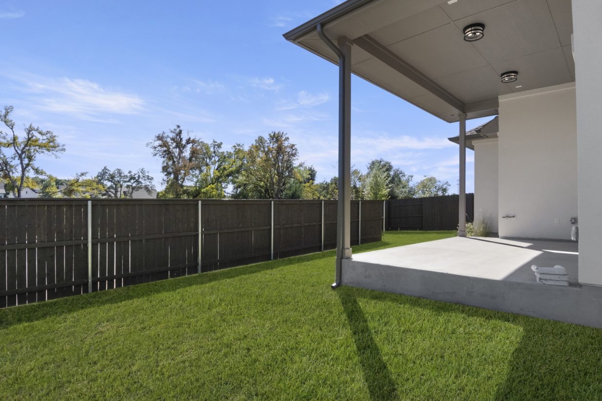 Image of a beige two story home exterior backyard with a covered stoop and a wooden fence, green grass, and a blue sky
