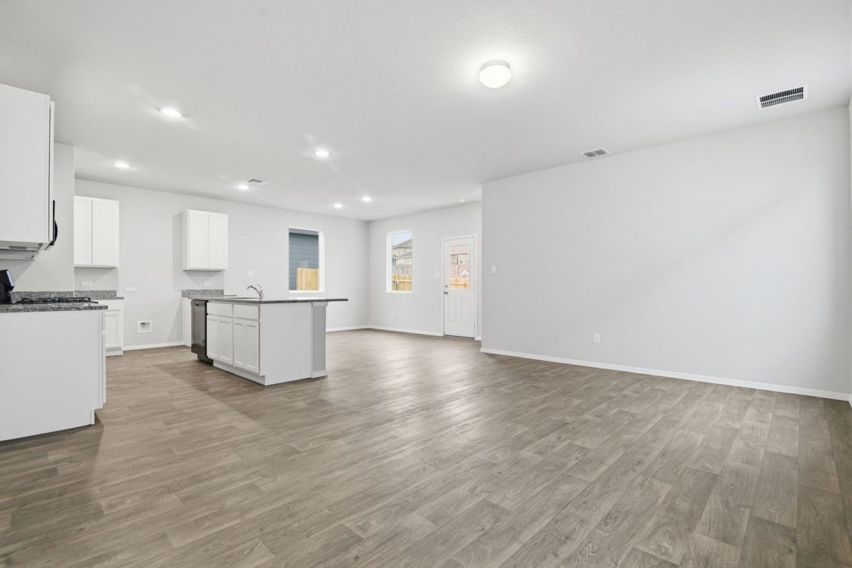 Image of a living room with light grey walls, brown flooring, an open kitchen, windows and white trim