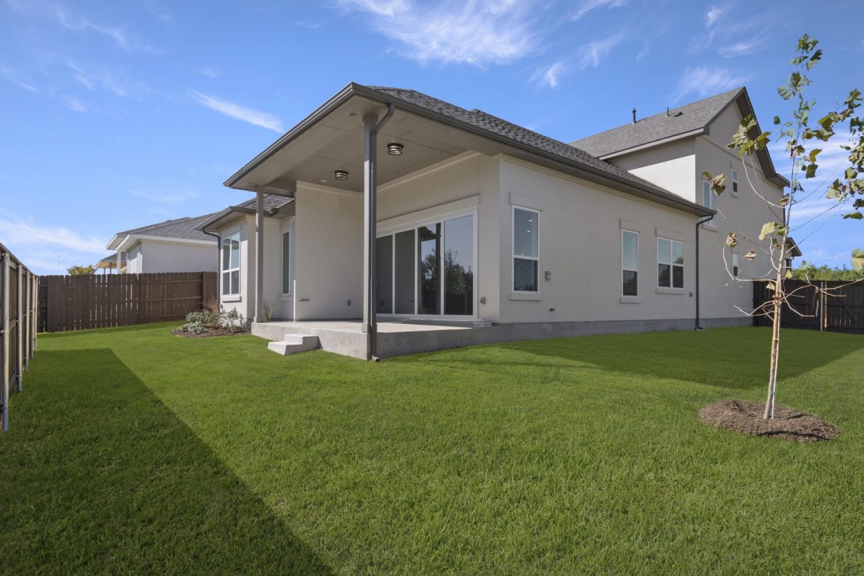 Image of a beige two story home back exterior with a glass door and green grass with a wooden fence and a blue sky