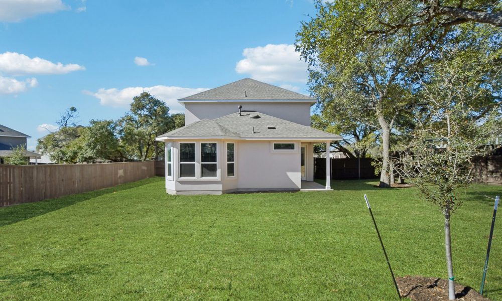 Image of the back exterior of a white two story home with a green grass back yard, a wooden fence, trees and a blue sky in the background