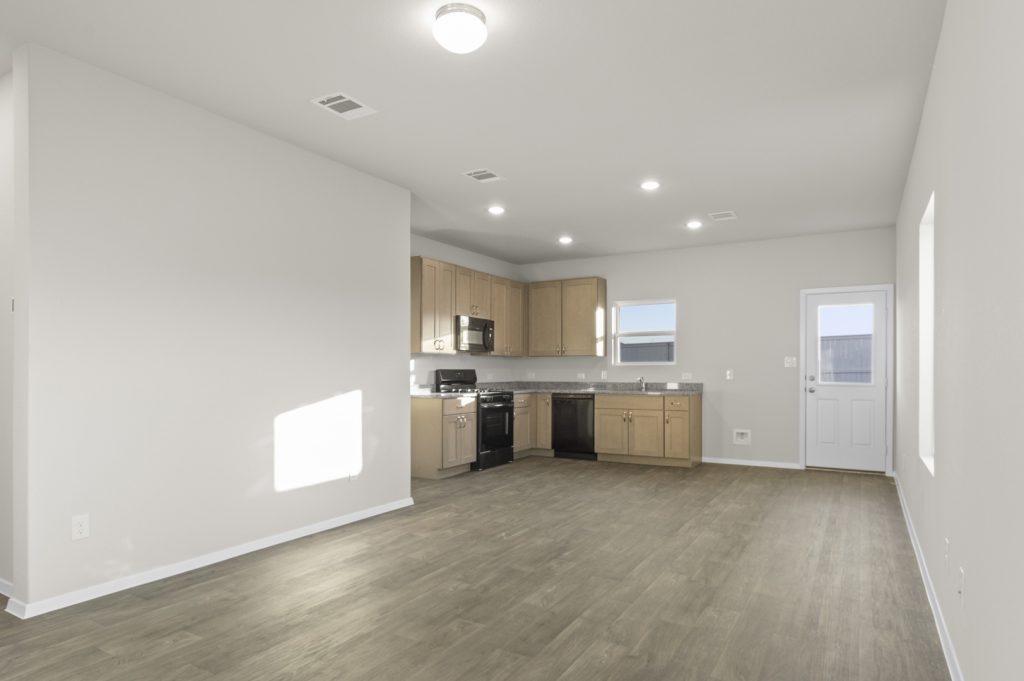 Image of one story home living and dining room area with dark wood-like flooring and light grey painted walls with a kitchen in the distance