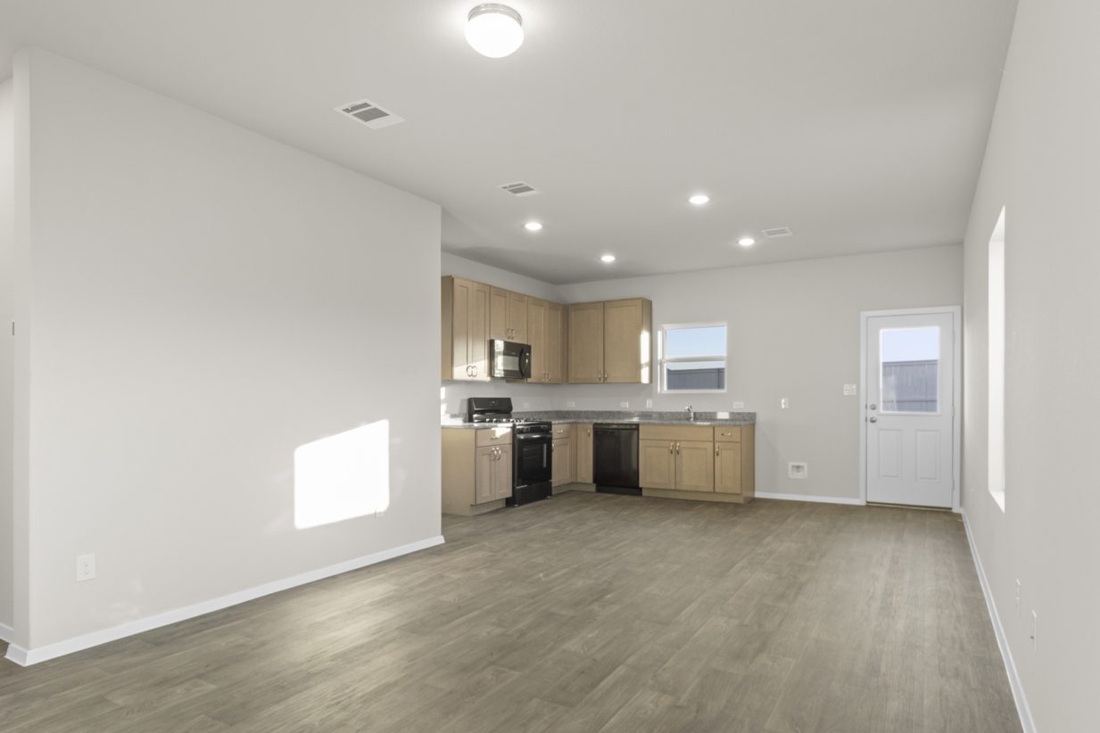 Image of one story home living and dining room area with dark wood-like flooring and light grey painted walls with a kitchen in the distance