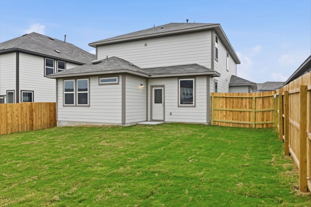Image of a light brown two story house exterior with brown trim, windows, a wooden fence, and a green grass backyard