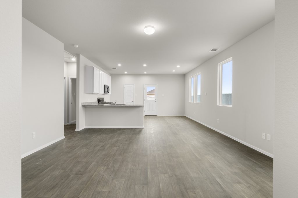 Image of a living room with cream walls, brown vinyl flooring, an open kitchen and windows
