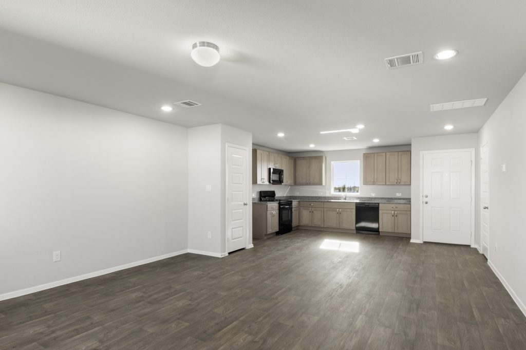 Image of a cottage home living room with dark vinyl flooring, light grey walls, and a L-shaped kitchen in the distance