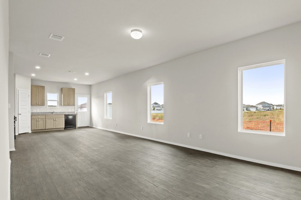 Image of a living room with brown flooring and light grey walls with windows and kitchen in the distance