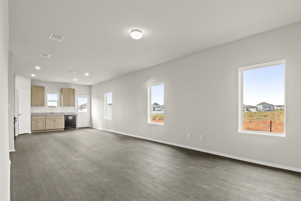 Image of a living room with brown flooring and light grey walls with windows and kitchen in the distance