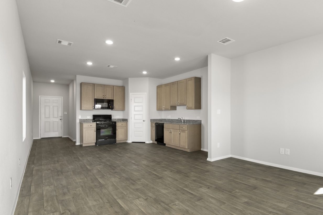 Image of a one story house living room with brown flooring and light grey walls with a kitchen in the distance