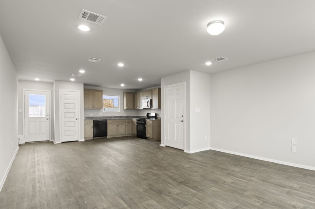 Image of a cottage home living room with brown vinyl flooring, light grey painted walls and a L-shaped kitchen in the distance