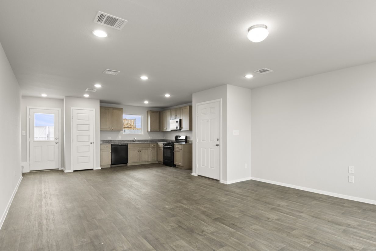 Image of a cottage home living room with brown vinyl flooring, light grey painted walls and a L-shaped kitchen in the distance