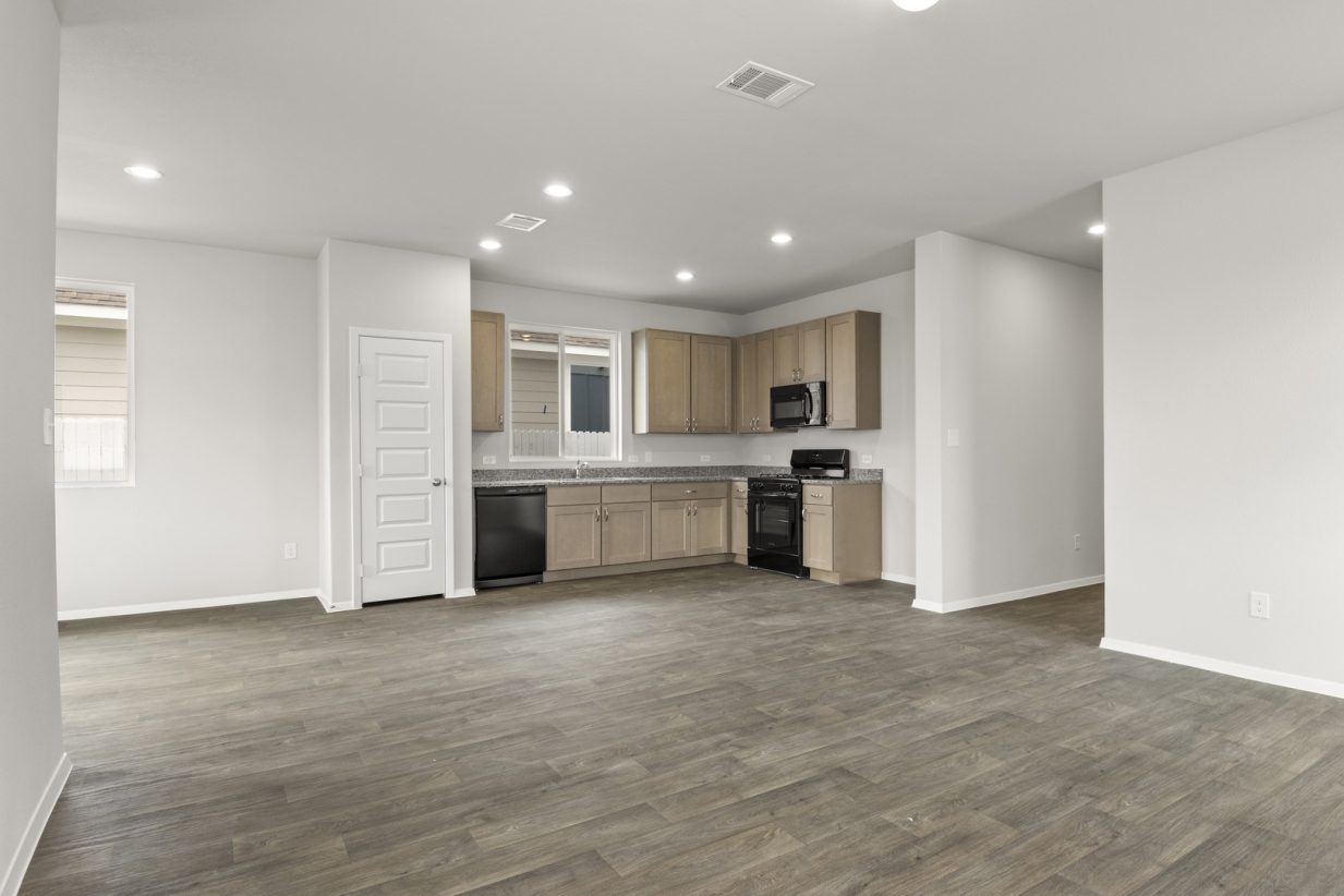 Image of two story home living room with brown flooring and light grey walls with a kitchen in the distance
