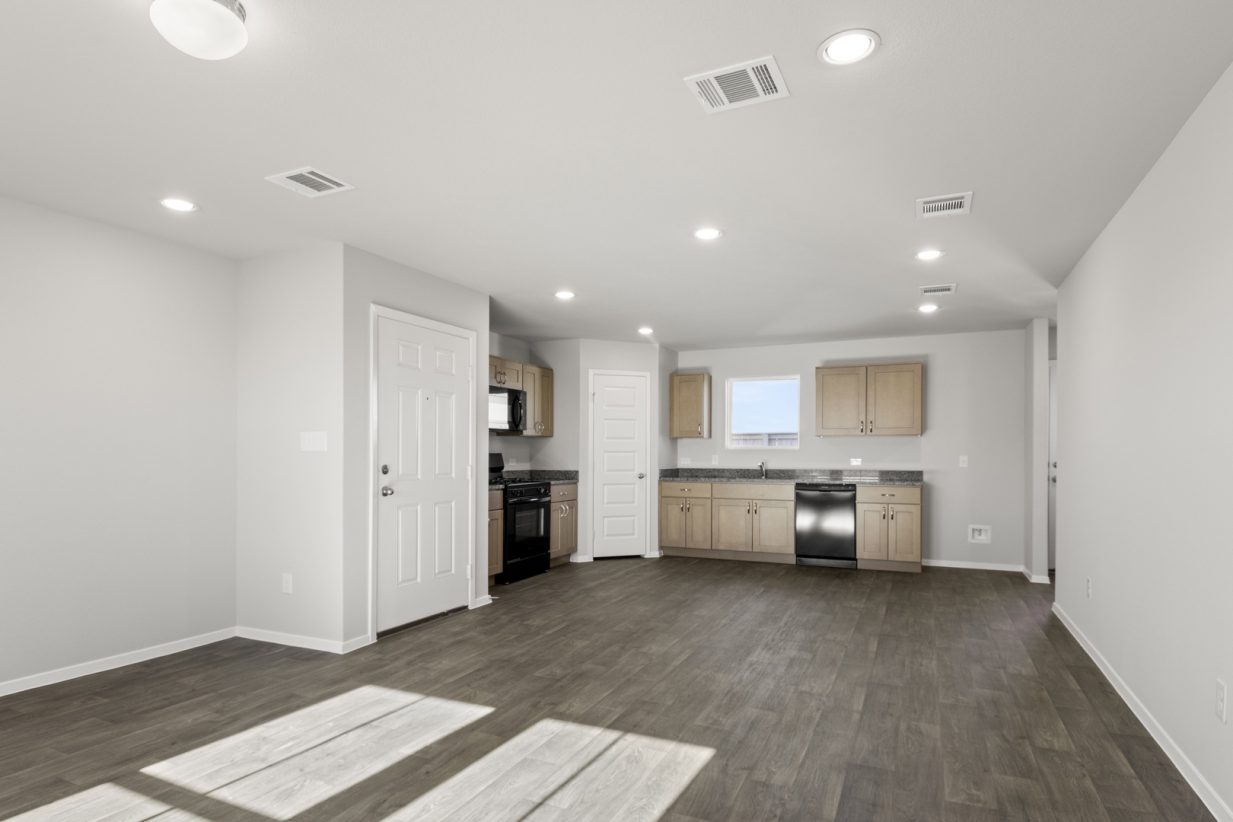 Image of a cottage home living room with dark vinyl flooring, light grey walls, and a L-shaped kitchen in the distance