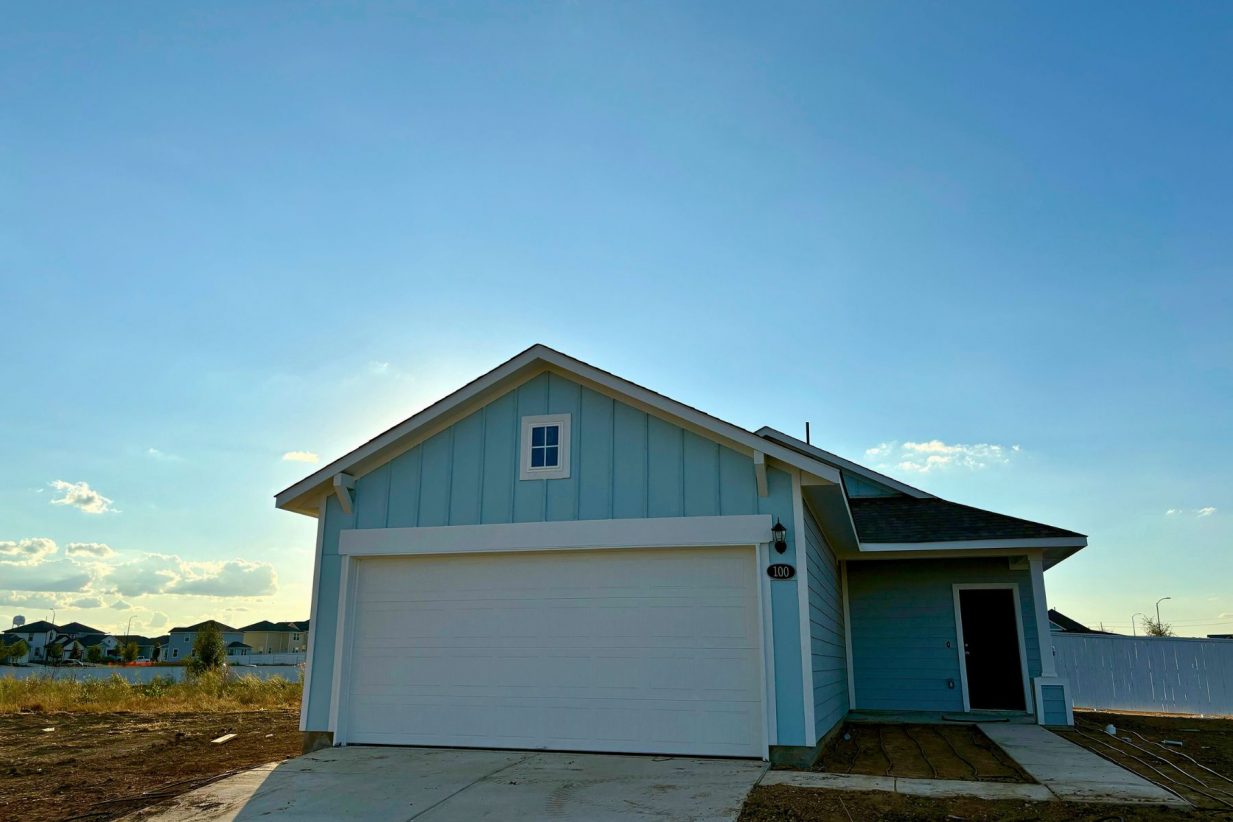 Image of one-story light blue home with blue door and white garage. Sidewalk from the driveway to the front door, dirt, and sky
