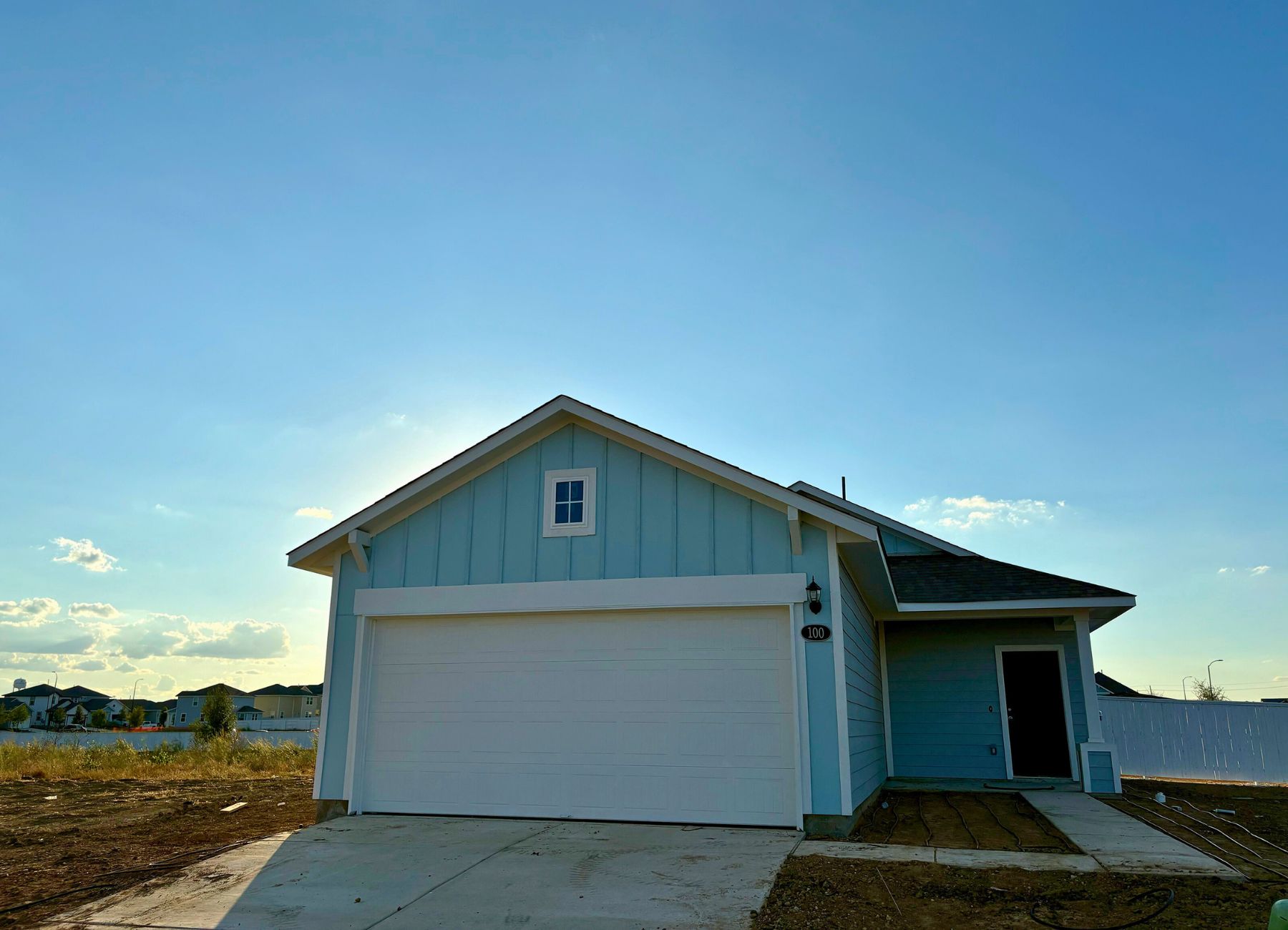 Image of one-story light blue home with blue door and white garage. Sidewalk from the driveway to the front door, dirt, and sky