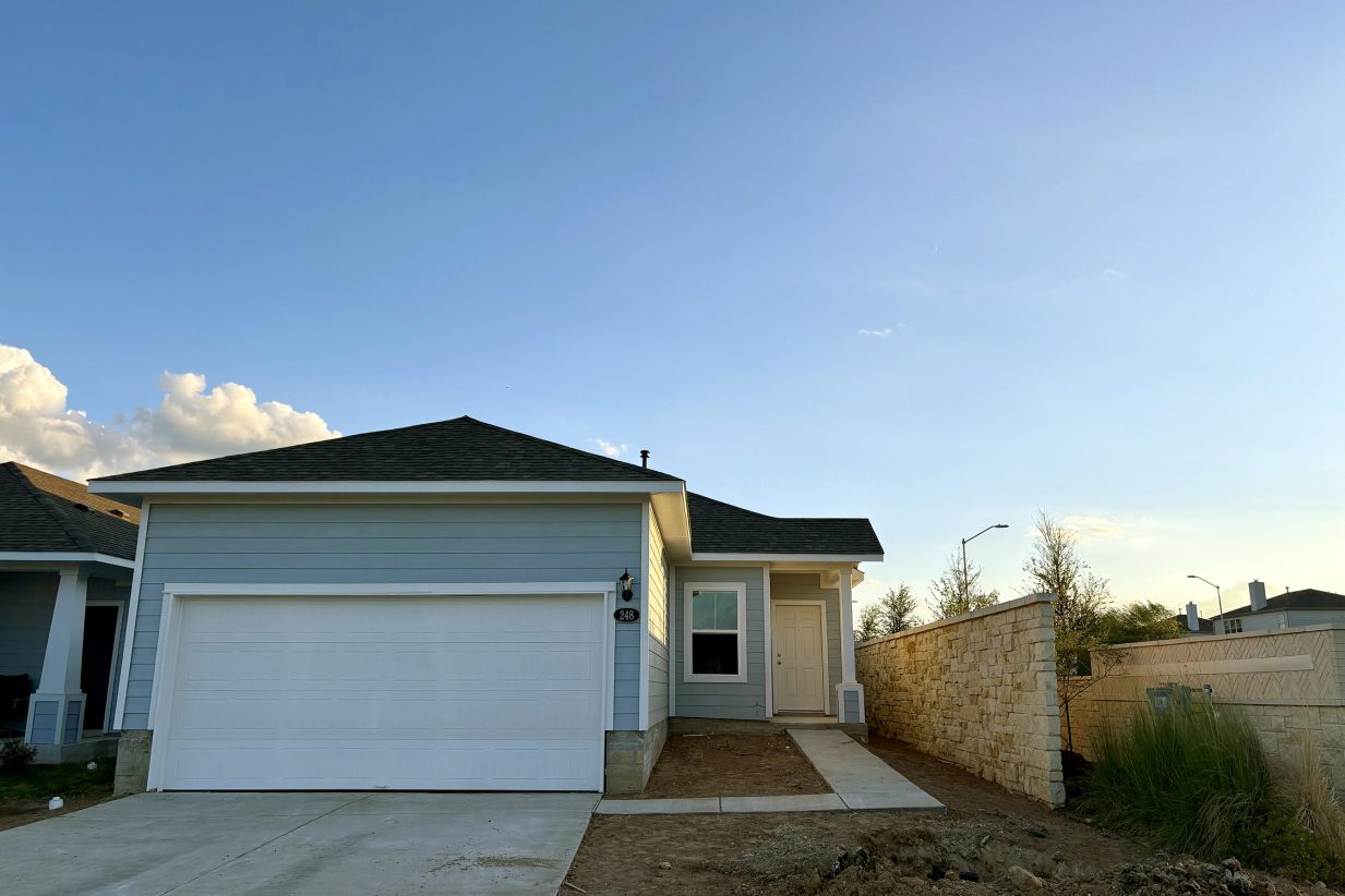 Image of one-story blue home with white trim and garage door with sidewalk from the driveway to the front door. Stone wall on one side of the home and sky