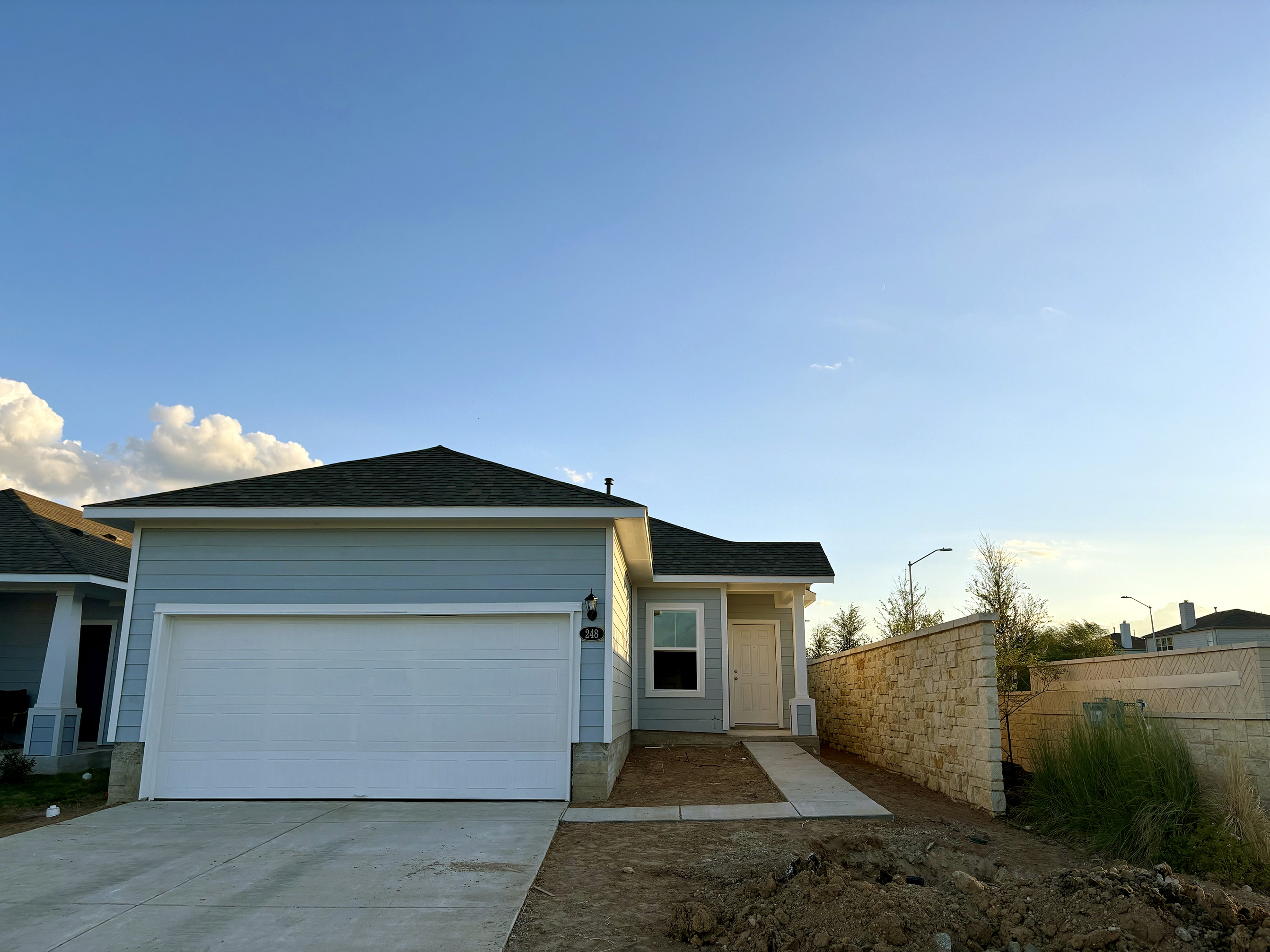 Image of one-story blue home with white trim and garage door with sidewalk from the driveway to the front door. Stone wall on one side of the home and sky
