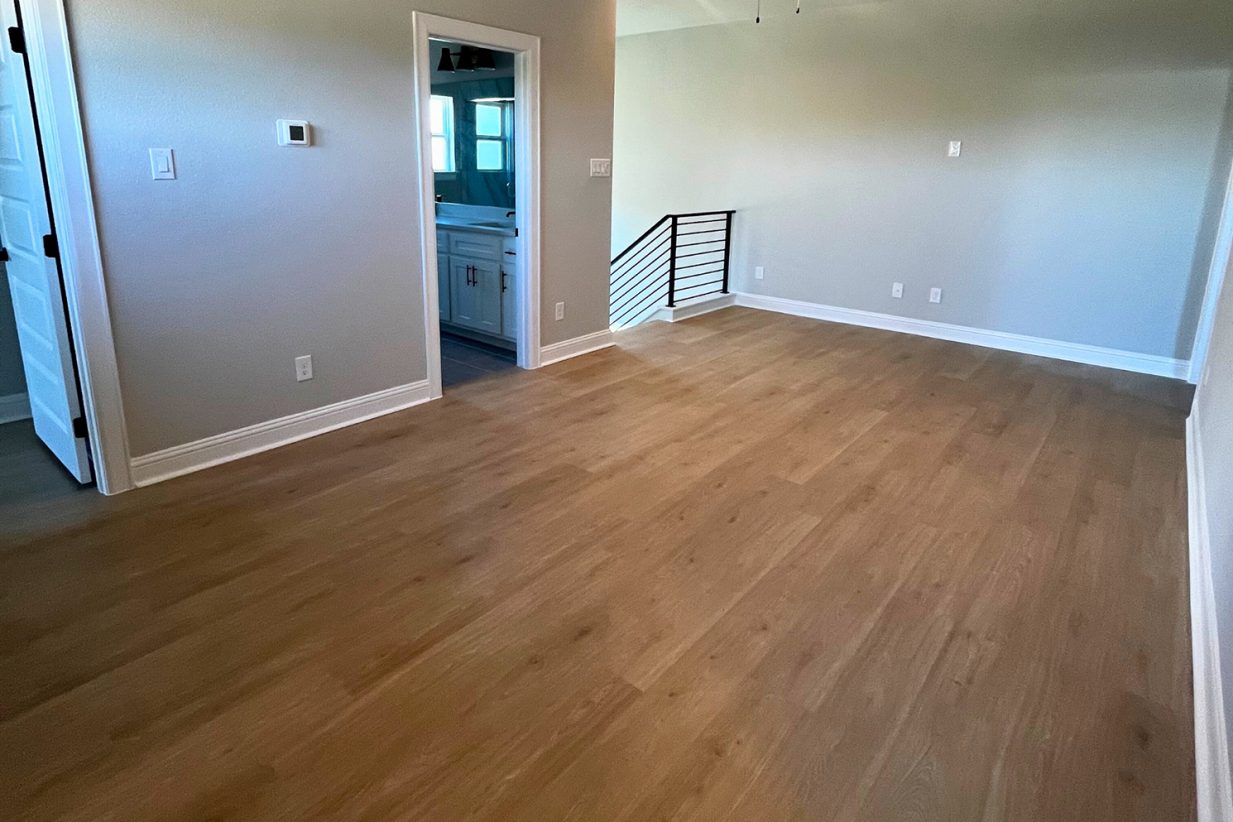 Image of the game room with wood flooring, gray painted walls, white base boards, and a black staircase railing leading downstairs.