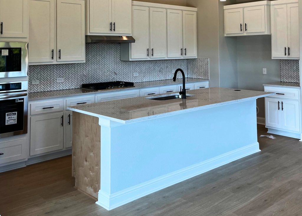 Image of kitchen with wood flooring, white cabinetry, stainless steel kitchen appliances, gray and white tiled blacksplash, a center island with white marbled countertop and a sink.