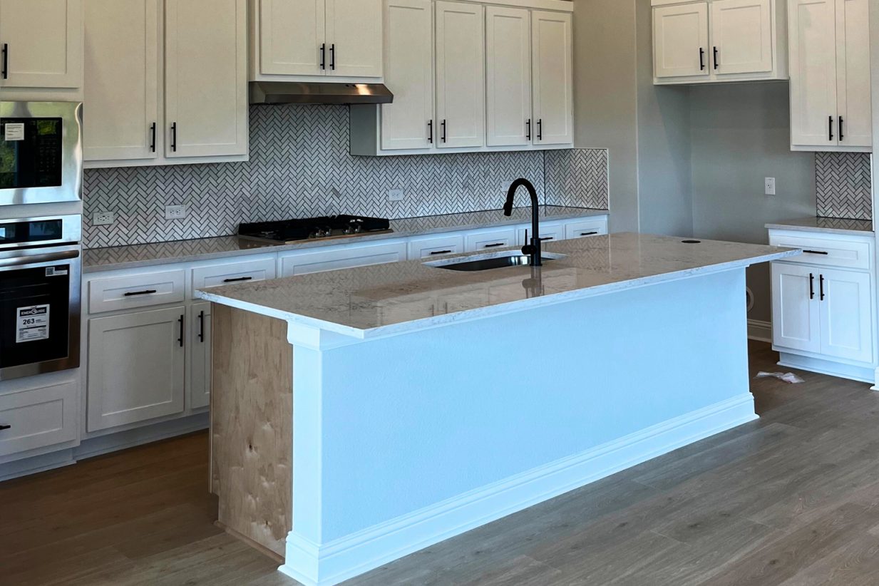 Image of kitchen with wood flooring, white cabinetry, stainless steel kitchen appliances, gray and white tiled blacksplash, a center island with white marbled countertop and a sink.