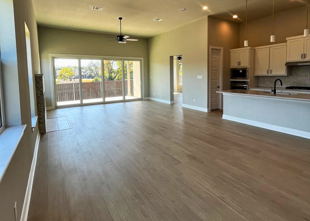 Image of living room and kitchen with wood flooring, with tan painted walls, brown ceiling fan, door windows, and a white countertop island and white cabinetry.