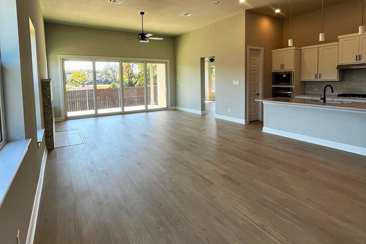 Image of living room and kitchen with wood flooring, with tan painted walls, brown ceiling fan, door windows, and a white countertop island and white cabinetry.
