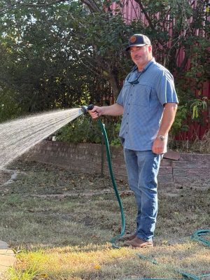 Image of a man wearing a blue shirt and jeans holding a green hose with water sprouting out of the hose. Grass an d trees are surrounding the man.