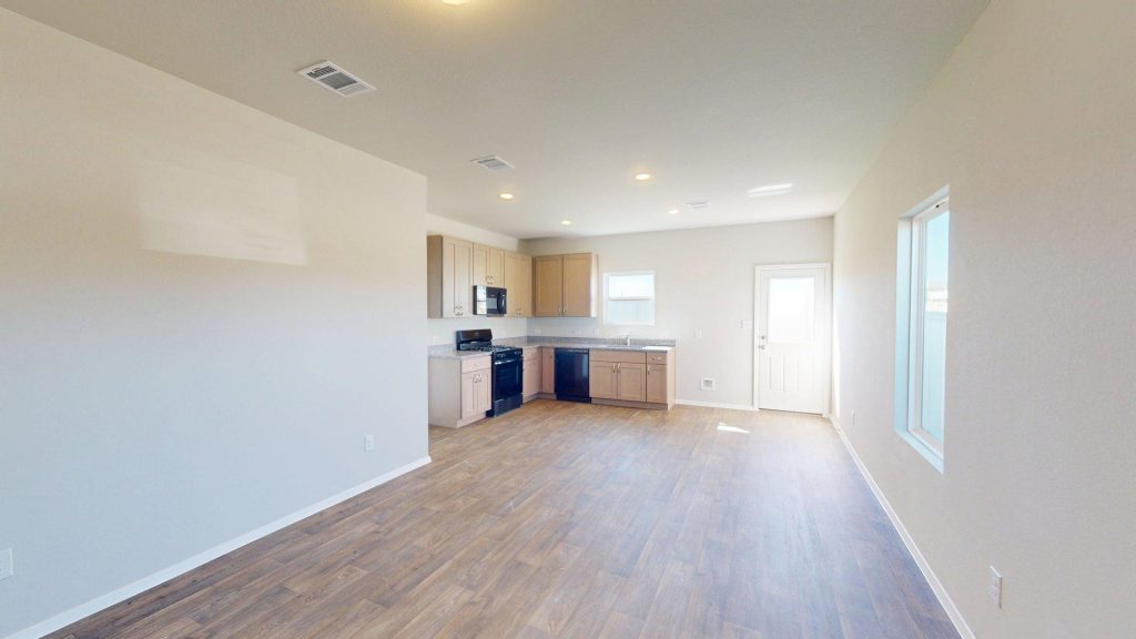 Image of one story home living and dining room area with dark wood-like flooring and light grey painted walls with a kitchen in the distance
