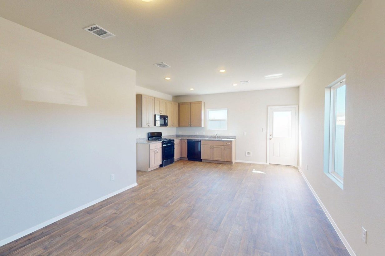 Image of one story home living and dining room area with dark wood-like flooring and light grey painted walls with a kitchen in the distance
