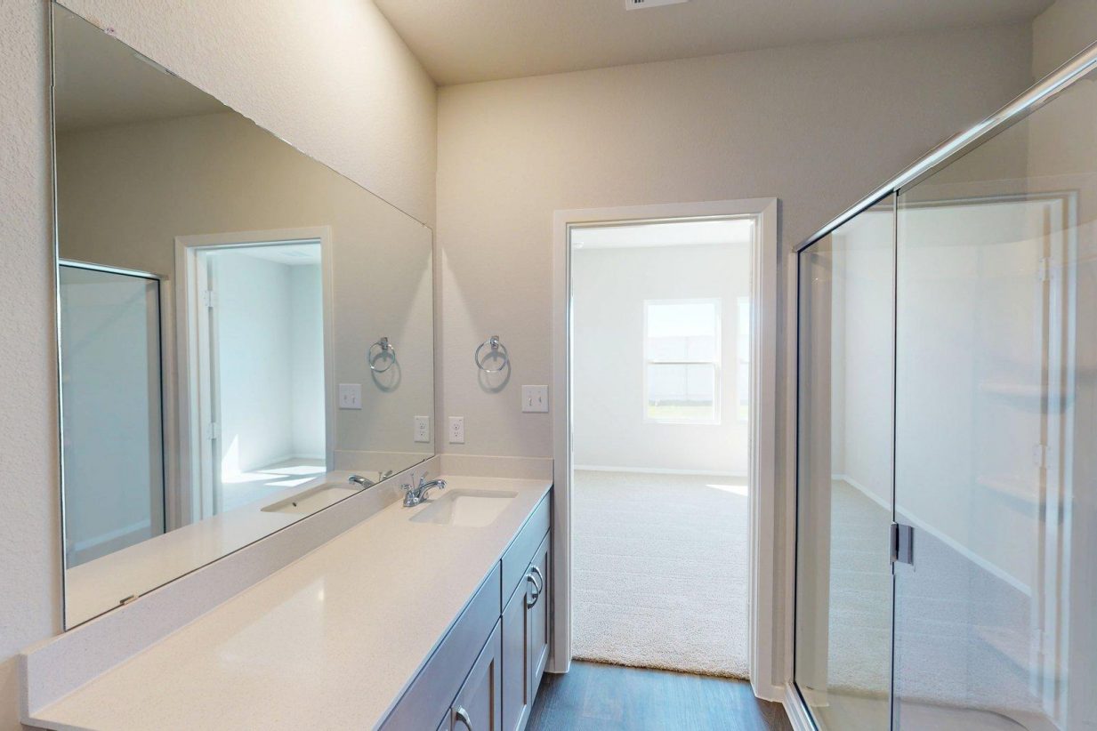 Image of a bathroom with a large mirror and white vanity with a walk-in shower and a door leading to the primary bedroom