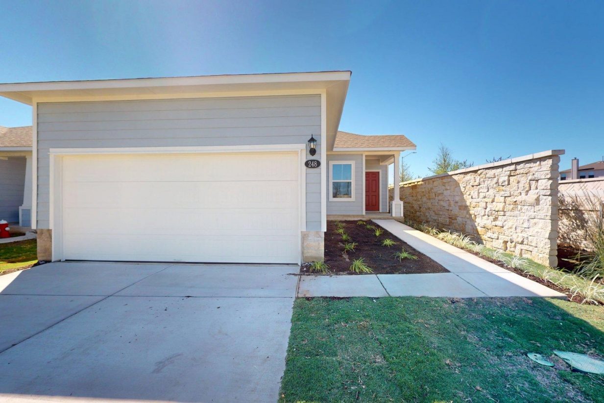 Image of exterior grey one story home with a red door and white two car garage with a cement driveway and a blue sky