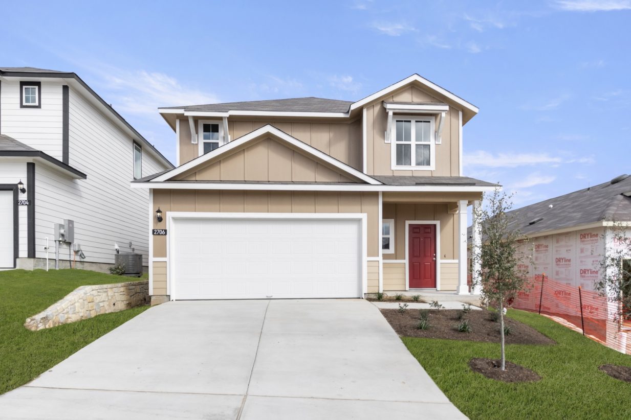 Image of the front exterior of a tan two story home with a white garage. red door, a cement driveway and a blue sky in the background