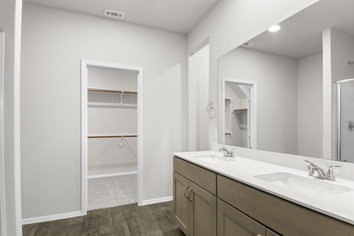 Image of a primary bathroom with brown cabinets, a white double vanity, a large mirror and a walk-in closet