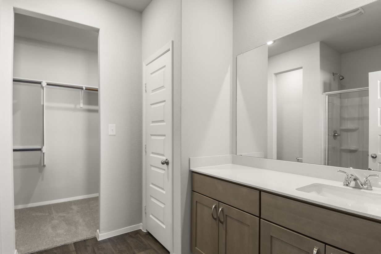 Image of a primary bathroom with brown cabinets, a white vanity, a large mirror and a walk-in closet