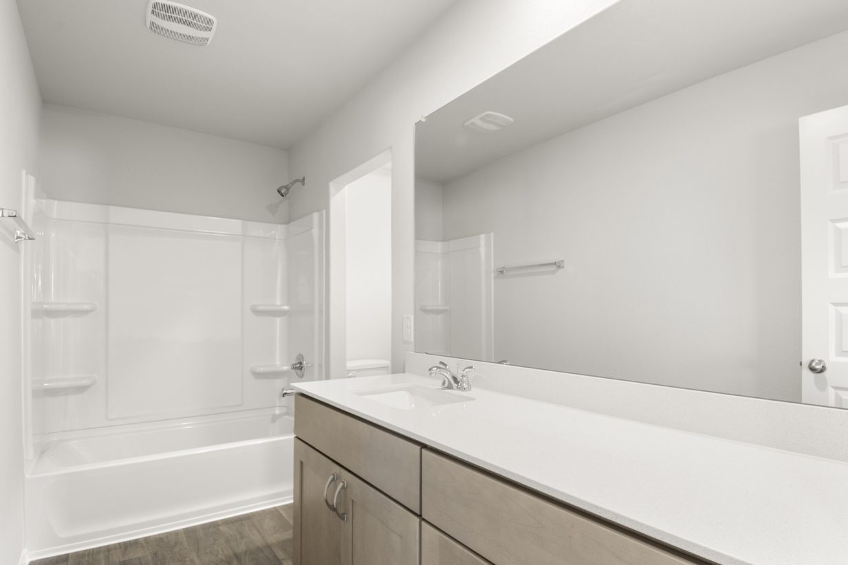 Image of a bathroom with brown cabinets and a white vanity with a white vanity and large mirror with white vinyl shower and tub