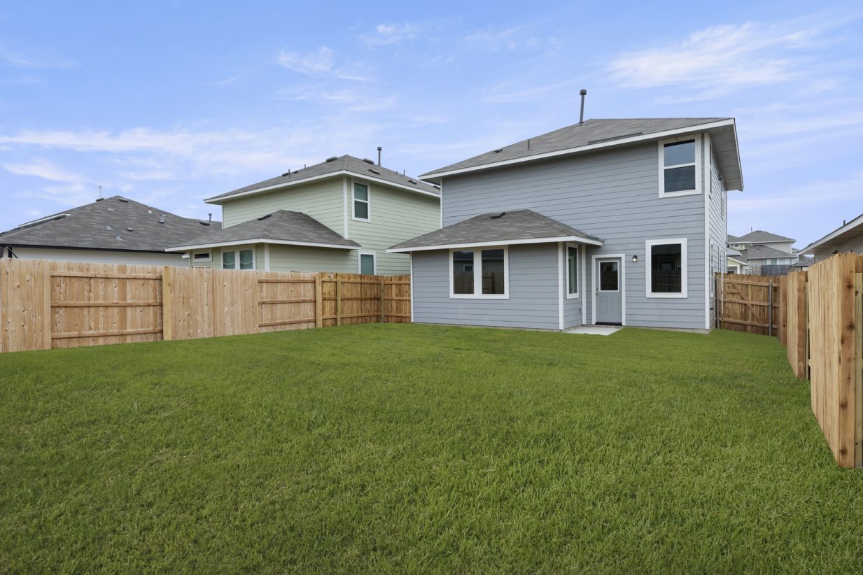 Image of a grey two story house back exterior with a large green grass backyard, a wooden fence, and a blue sky in the background